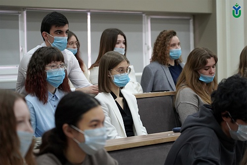 На изображении запечатлена группа людей, сидящих рядами, по всей видимости, в аудитории или конференц-зале. Большинство людей носят синие медицинские маски, закрывающие нос и рот.  В первом ряду слева видна девушка с распущенными каштановыми волосами, смо