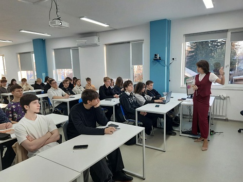 В классе идёт урок. Ученики сидят за партами, а учительница стоит перед ними и что-то объясняет, жестикулируя правой рукой. Учительница одета в белый верх и бордовый костюм-тройку (жилет и брюки).  Ученики внимательно смотрят на учительницу, некоторые из 