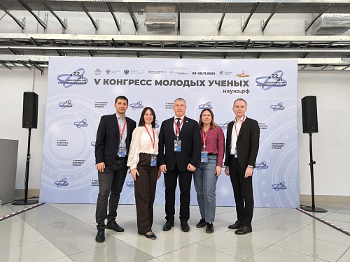 На фотографии пять человек, стоящих на фоне пресс-волла. Пресс-волл белого цвета с логотипом и надписями. Слева и справа от пресс-волла находятся черные колонки. Пол покрыт белыми плитками с серым рисунком.  На пресс-волле изображен логотип с надписью 