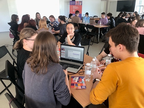 На изображении представлен интерьер помещения, в котором проходит мероприятие с участием большого количества людей. Они сгруппированы вокруг столов и занимаются общей деятельностью.  На переднем плане группа из четырех человек работает за столом. На столе