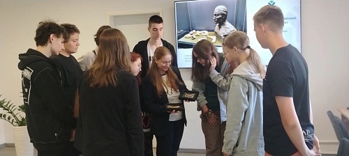 Изображение показывает группу людей, стоящих в помещении. В центре кадра две женщины держат небольшие рамки, в которых находятся предметы, напоминающие костяные или керамические осколки, расположенные в линию. Остальные люди, стоящие вокруг, смотрят на эт