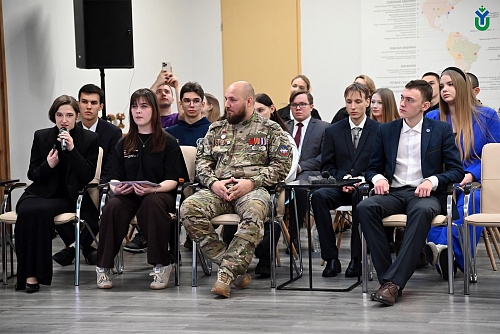 На фотографии запечатлена группа людей, сидящих в ряд на стульях в помещении. Стулья расположены близко друг к другу, и люди, сидящие на них, обращены лицом к зрителю.  В первом ряду слева направо: девушка в черном костюме держит микрофон, говоря в него;