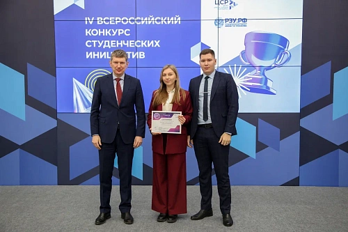 Три человека стоят на фоне баннера. Слева стоит мужчина в тёмно-синем костюме и красном галстуке. В центре - девушка в костюме бордового цвета с белой блузкой, держащая в руках сертификат. Справа находится мужчина в тёмно-синем костюме и тёмном галстуке.