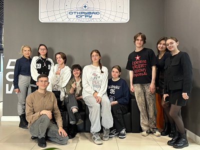 На фотографии группа из одиннадцати человек. Они стоят и сидят на диване на фоне серой стены. Над ними расположен прямоугольный знак с надписью 