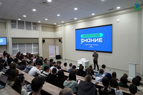 В аудитории находится большое количество людей, сидящих за рядами столов. Большинство из них обращены лицом к сцене, на которой стоит мужчина с микрофоном. На стене за ним расположен большой экран, на котором отображается текст: «Обеспечивая технологическ