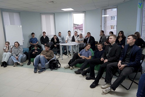 На фотографии запечатлена комната, в которой находится группа студентов, сидящих на пуфах и стульях.Слева у стены расположились две девушки в очках, сидящие на серых пуфах. Рядом с ними сидит молодой человек, смотрящий в телефон. Далее в кадре виден парен