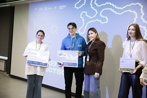 На фотографии четыре человека стоят в помещении перед экраном проектора.Слева направо: Женщина в белой футболке, свободных джинсах, в очках, с небрежно собранными волосами. Она держит в руках диплом, на котором написано 