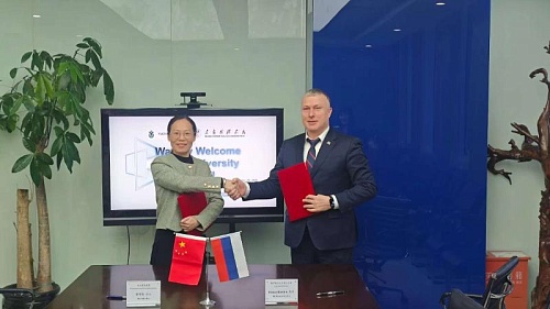 На изображении запечатлены два человека, пожимающие друг другу руки на фоне экрана телевизора. Женщина, стоящая слева, держит в руках красную папку. Мужчина справа, также держит в руках красную папку. Между ними, на столе, расположены флаги Китая и России