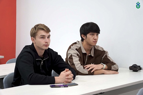 На фотографии запечатлены два молодых человека, сидящих за белым столом в помещении. Слева сидит парень со светлыми волосами в черной кофте на молнии, его руки сложены вместе. Справа от него сидит парень с темными волосами в коричнево-бежевой кофте на мол