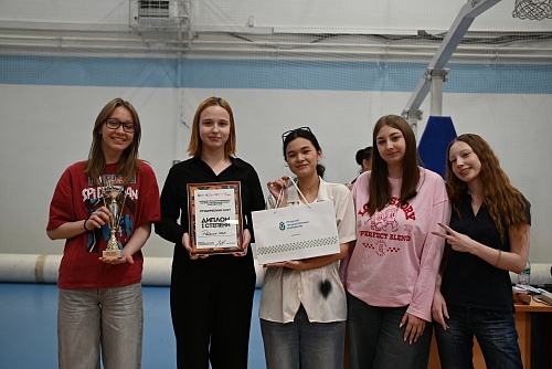 Пять девушек стоят в ряд, улыбаясь и позируя для фотографии. Первая слева девушка носит очки, красную футболку с рисунком Человека-паука, серые брюки и держит золотой трофей. Вторая девушка в черной рубашке и брюках держит в руках рамку с дипломом. На дип