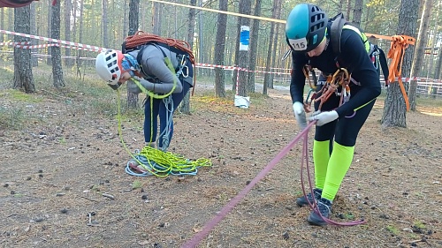 На фотографии изображены два человека, одетые в альпинистское снаряжение, в лесу. Первый человек слева носит белый шлем, серую куртку и синие брюки. У него на спине закреплена оранжевая веревка, а перед ним на земле лежит куча зелено-голубой веревки. Втор