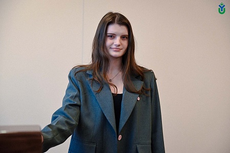 На изображении представлена молодая женщина с длинными каштановыми волосами, которые распущены и ниспадают на плечи. У нее светлая кожа, голубые глаза и легкая улыбка. На шее у нее висит тонкая цепочка с кулоном в форме сердца.  Она одета в темно-серый пи