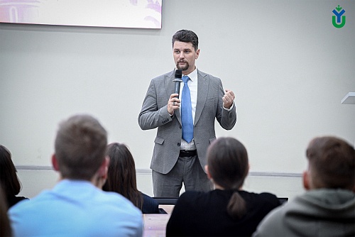 На изображении представлен мужчина, выступающий перед аудиторией. Он одет в серый костюм, белую рубашку и голубой галстук. Он держит микрофон в руке и жестикулирует, видимо, что-то объясняя.Аудитория состоит из нескольких человек, сидящих на стульях. Видн