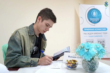 На фотографии изображен молодой человек, заполняющий документы за столом. На нем надета джинсовая куртка и черная футболка с принтом. В левой руке он держит развернутый паспорт в обложке, а правой руке — синюю шариковую ручку, которой он заполняет бланк.