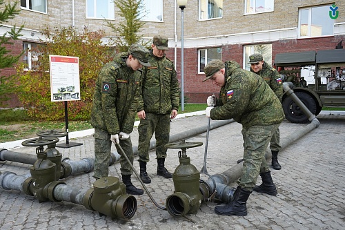 Четыре человека в военной форме занимаются соединением водопроводных труб. На земле лежат три больших вентиля, соединенных трубами. Один человек использует рычаг для затягивания соединения. Все четверо одеты в камуфляжную форму и черные ботинки. На рукава