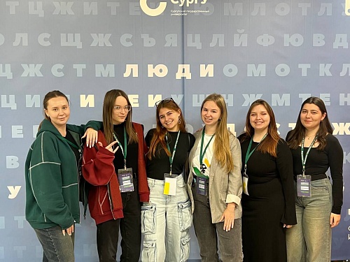 Шесть студенток ЮГУ стоят на фоне пресс-волла Регионального слёта волонтёров финансового просвещения Югры «Школа лидеров»