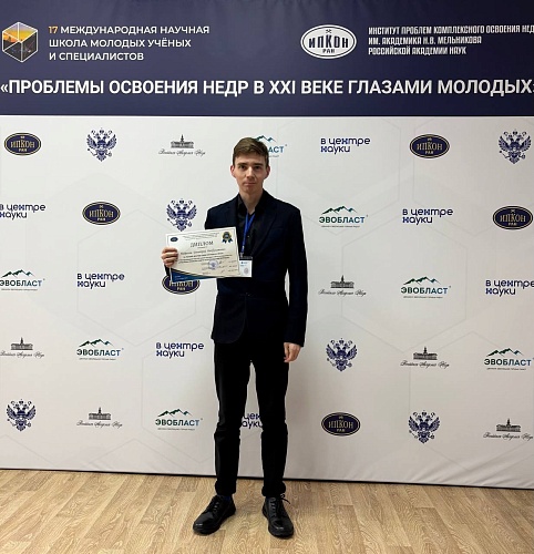 На фотографии запечатлен молодой человек, стоящий на фоне баннера с надписями и логотипами. Парень одет в черный костюм и черные туфли. В руках он держит диплом в рамке. На шее у него висит бейдж.На заднем плане находится баннер, на котором указана информ