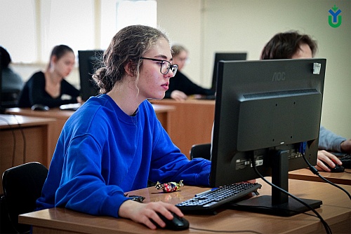 На изображении показан компьютерный класс. На переднем плане сидит девушка в синей толстовке и очках, работающая за компьютером с монитором AOC. Рядом с клавиатурой лежит конфета в обертке. Девушка смотрит на монитор, ее руки лежат на мышке.В глубине клас