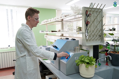 На фотографии изображен мужчина в белом лабораторном халате и прозрачных защитных очках, который стоит у лабораторного стола и управляет устройством. Устройство имеет серый корпус с синей панелью управления, на которой расположены кнопки красного и зелено