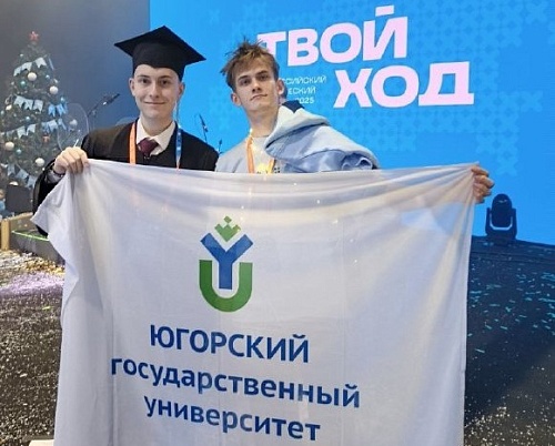 На фотографии два молодых человека держат флаг Югорского государственного университета. На флаге изображен логотип университета, а также надпись 