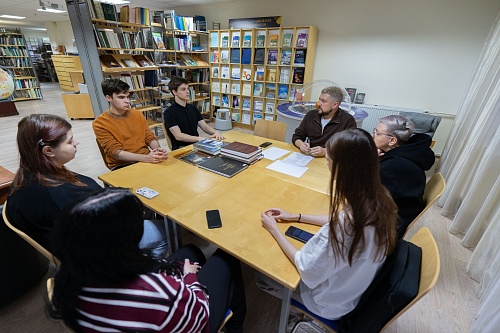 В помещении музея за большим прямоугольным столом собралась группа из семи человек. Стол состоит из нескольких соединённых частей и на нём лежат стопки книг, бумаги и мобильные телефоны.Слева сидит девушка с тёмными волосами, рядом с ней - молодой человек