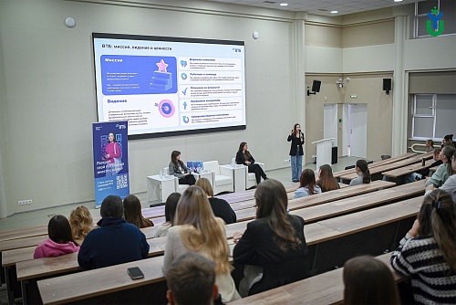 На снимке представлен интерьер актового зала или лекционной аудитории. На переднем плане находятся ряды деревянных парт, заполненные людьми, сидящими спиной к камере. В центре кадра, перед аудиторией, располагается небольшая сцена. На сцене установлены тр