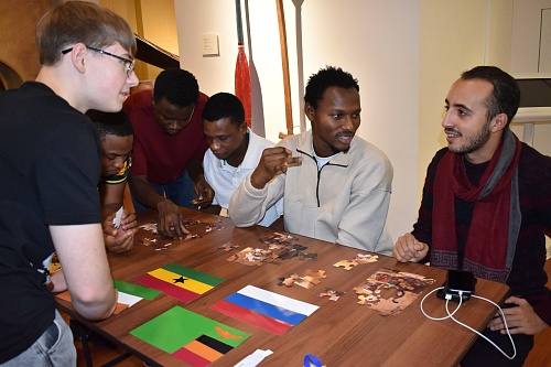 На фотографии группа из шести человек, собравшихся вокруг деревянного стола, чтобы собрать пазл. На столе, помимо разбросанных частей пазла, лежат три маленьких флага: Ганы, России и Замбии.В центре стола несколько людей собирают части пазла. Один из них 