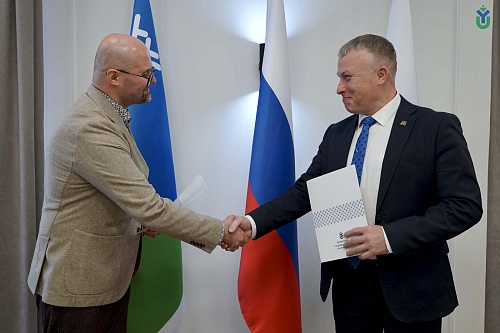 Ректор ЮГУ Роман Кучин и руководитель академического направления Фонда региональных социальных программ «Наше будущее» Пётр Сафронов поживают руки на фоне флагов России, Югры и ЮГУ в знак подписания соглашения