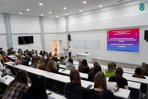 Изображение показывает аудиторию, заполненную студентами, сидящими за партами в лекционном зале. В передних рядах в основном видны затылки и прически. Впереди стоит лектор у трибуны, произносящий речь.  На стене позади лектора висит большой экран, отображ