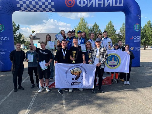 Группа студентов в том числе ЮГУ стоит на пьедестале на открытом воздухе, держат трофеи, дипломы чемпионата по спортивному ориентирвоанию и два флага с надписью ЮГОРСКИЙ ГОСУДАРСТВЕННЫЙ УНИВЕРСИТЕТ и СЕВЕР