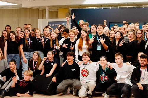 На фотографии изображена большая группа людей, собравшихся вместе. Многие из них одеты в темную одежду и носят медали на лентах цветов российского флага. На некоторых людях надеты бейджи с логотипом. Некоторые участники группы показывают различные жесты р