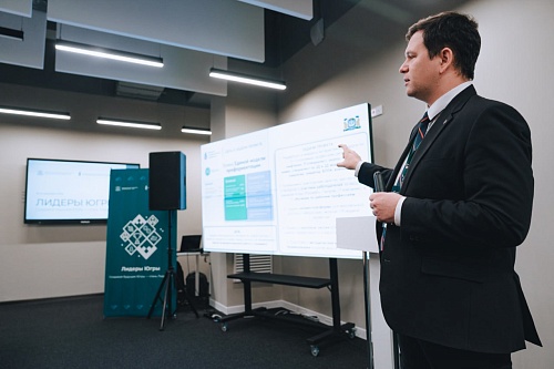 На изображении показан мужчина в черном костюме и галстуке, стоящий справа от интерактивной доски. Он указывает пальцем на доску и держит в руке листок бумаги. На доске отображена презентация, содержащая текст и графики. Виден заголовок: 