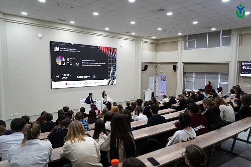 Изображение показывает аудиторию, сидящую рядами деревянных скамеек с прикрепленными столами. В передней части аудитории на сцене находятся два человека, сидящие в белых креслах, и между ними стоит маленький столик. На заднем плане за ними висит большой э