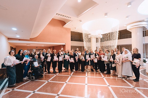 В просторном зале с высоким потолком группа людей позирует для фотографии. Пол выложен плиткой терракотового цвета с белыми вставками. В центре потолка висит большая круглая люстра, состоящая из множества светящихся нитей.  Люди стоят в два ряда. Многие д