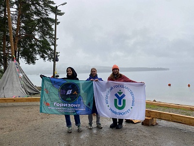 Три студентки ЮГУ стоят на берегу озера Тургояк в Челябинской области, держа в руках флаги с эмблемой ЮГУ и турклуба «Горизонт»