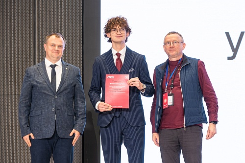 На фотографии изображены три человека, стоящие на фоне стены с вертикальными полосами слева и белого фона с большой серой буквой 