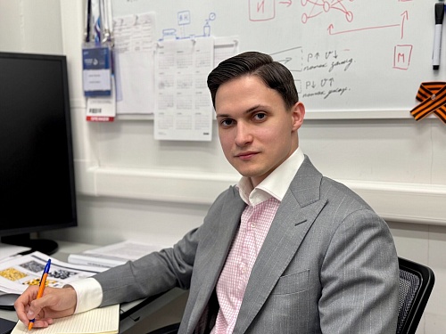 Мужчина в сером костюме сидит за столом. У него короткая темная прическа, светлая кожа и внимательный взгляд. Он одет в белую рубашку с розовым узором и серый пиджак в тонкую полоску. В правой руке у него оранжевая ручка, и он что-то записывает в блокнот,