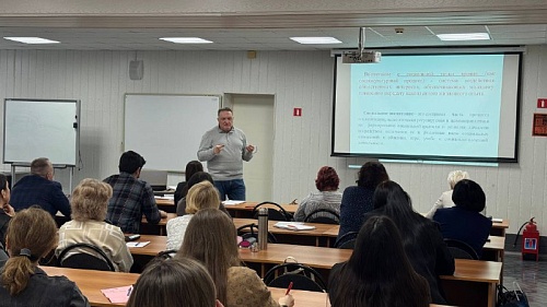 Изображение показывает аудиторию, в которой проходит лекция или семинар. Комната хорошо освещена, вероятно, люминесцентными лампами на потолке. В передней части комнаты стоит мужчина, который, судя по всему, является лектором. Он одет в серый свитер и син