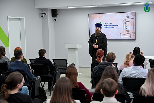 Изображение показывает аудиторию студентов, сидящих на черных креслах и смотрящих на священника в черной рясе с золотым крестом на шее, стоящего перед белой трибуной.На заднем плане висит экран, на котором отображено название 