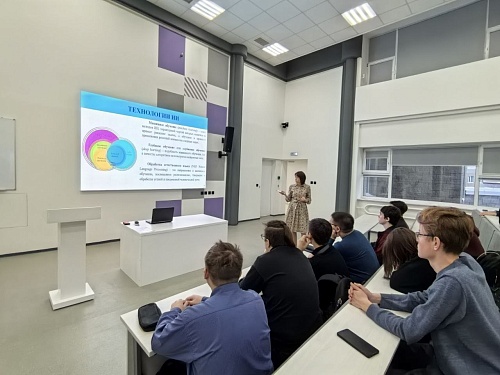 Изображение показывает лекционную аудиторию. В передней части комнаты, слева, стоит белая трибуна. На трибуне расположен ноутбук и несколько листов бумаги. За трибуной находится белая тумба, на которой также лежит ноутбук и бумага. На стене висит большой 