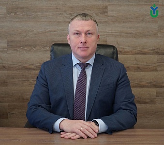 На изображении мужчина, сидящий за столом. Он одет в темно-синий костюм, голубую рубашку и галстук с темно-бордовым узором. Его руки сложены на столе, на безымянном пальце правой руки надето золотое кольцо. У мужчины короткая стрижка, светлые волосы и гол