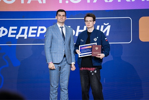 На изображении два человека стоят на фоне синего баннера с белыми графическими элементами и частично видимым текстом. Верхняя часть баннера окрашена в персиковый цвет.Слева стоит мужчина в голубом клетчатом костюме, белой рубашке и темно-синем галстуке. Н