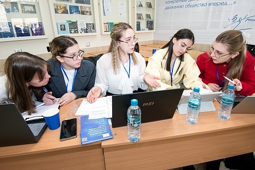 Пять девушек сидят за одним столом и, по-видимому, работают над проектом. На столе стоят ноутбуки, планшет, бутылки с водой «Черноголовка», телефон, синий стакан, бумаги и ручки. На девушках надеты блузки и жакеты, у всех на шее бейджики с синей лентой.На