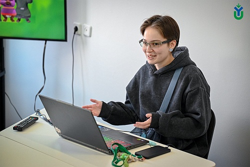 На изображении девушка с короткими волосами, в очках и в темной толстовке с капюшоном. У неё через плечо перекинута светло-синяя сумка. Она сидит за белым столом перед открытым ноутбуком и активно жестикулирует руками, как будто разговаривает с кем-то.  Н